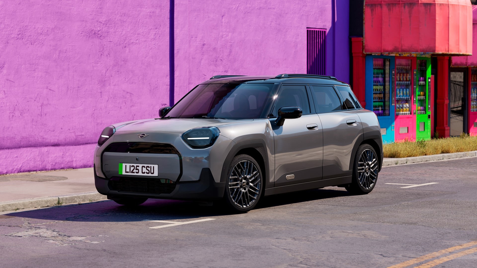 Gray MINI Aceman parked on a city street beside a bright purple and red building, featuring modern design and black roof.